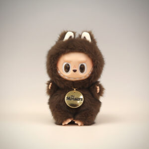 A brown, fuzzy "The Monsters Little Leader" doll with white-lined ears, a smiling light-skinned face, and a golden "The Monsters" medallion around its neck, seated on a smooth, light gray to off-white gradient background.