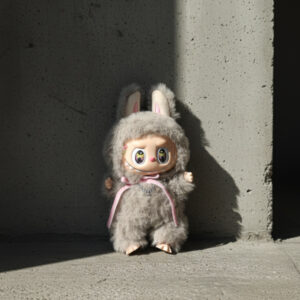 A grey, fluffy designer toy doll with large expressive eyes, a pink bow, and bunny ears, standing in a strip of sunlight against a rough concrete wall, casting a strong shadow.
