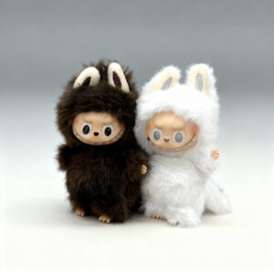 Two collectible fuzzy plush dolls standing side-by-side: one dark brown and one white. Both figures have vinyl faces with mischievous smiles, large eyes, and furry hoods with ears. They are centered on a clean white-to-gray gradient background.