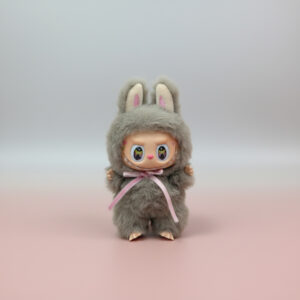 A cute, fuzzy gray plush doll wearing a bunny hood with pink inner ears, featuring a smiling vinyl face with large, sparkly eyes and a pink nose, tied with a delicate pink ribbon, standing on a soft pink and gray gradient background.