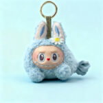 Close-up of a light blue, fuzzy plush keychain with bunny ears, a smiling doll-like face with a heart nose and large eyes, and a small white daisy accessory, displayed on a matching light blue background.