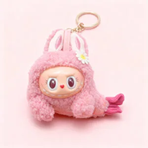 Close-up of a pink, fuzzy plush keychain with bunny ears, a smiling doll-like face with a heart nose and large eyes, and a small white daisy accessory, displayed on a matching pink background.